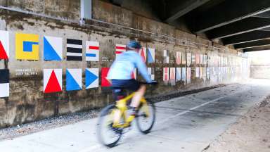 A biker whizzes by the Alluvial Decoder Floodplain educational mural that is the name of significant storms spelled out using nautical flags