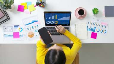 arial photo of woman looking at her computer with a screen that says "business analysis." There are pages of reports on the table around the computer.