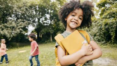 smiling child holding school books with friends playing in the background
