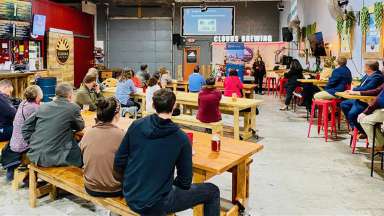 A bar type area with people sitting at tables listening to a person speak. A TV shows presentation in background.