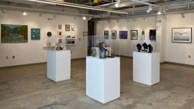 A wide view of Pullen Arts Center's Gallery with white walls and concrete floors. There are four white pedestals in center with ceramics and jewelry on display. There is two dimensional hanging on the walls as well. 