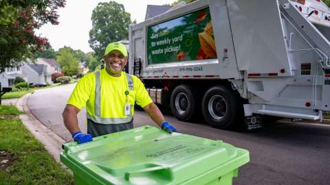 A Solid Waste Collector stands by a yard waste bin and trash truck.