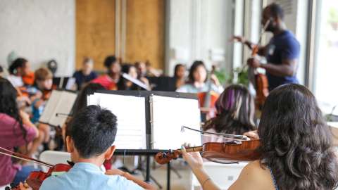 A group of students play string instruments with a person leading them.