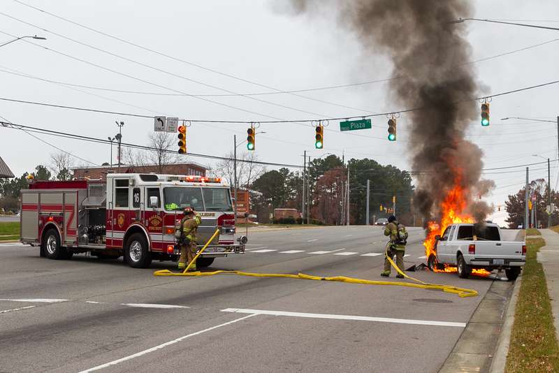 Two firefighters operate a fire hose on a car engulfed in flames