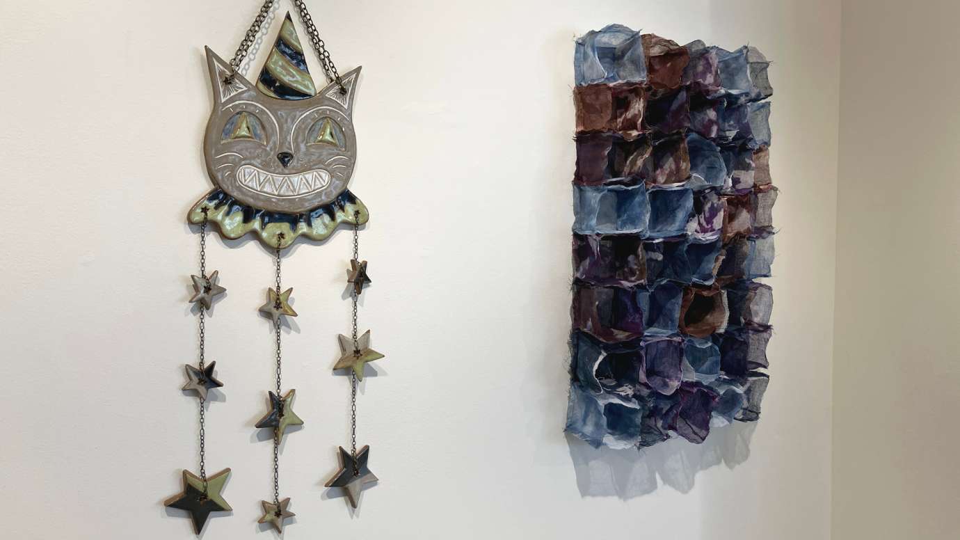 Two works hung on the gallery wall: one a ceramic work of a smiling cat in a hat with strands of stars, and one three-dimensional fiber work in shades of blue, red and purple
