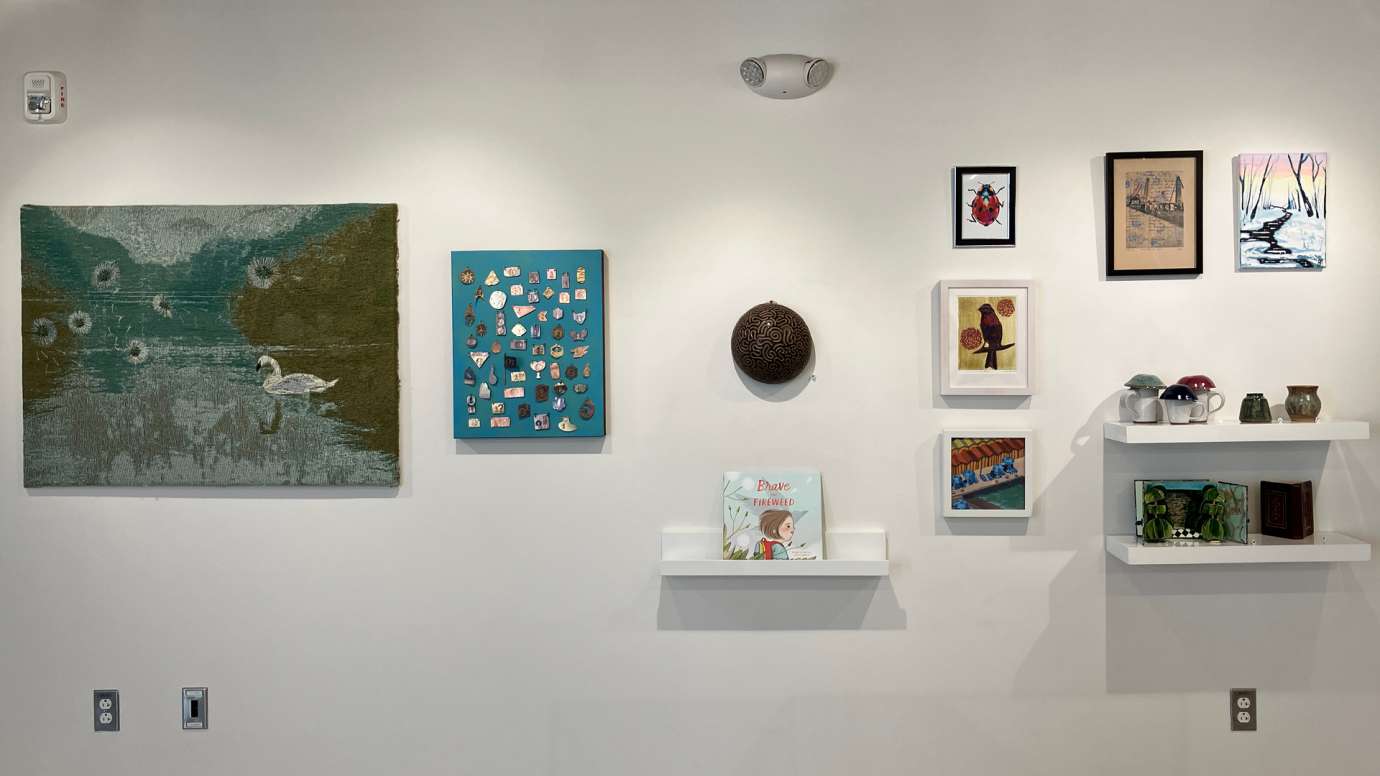 Installation shot of the gallery with a mixture of two and three dimensional works shown, in various media including painting, pottery, an illustrated children's book, jewelry, fiber works, and more
