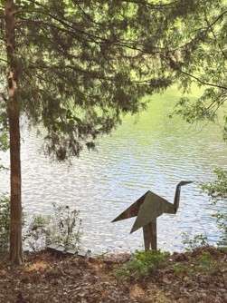 Silouette of a folded metal Heron at the side of a wooded lake. 