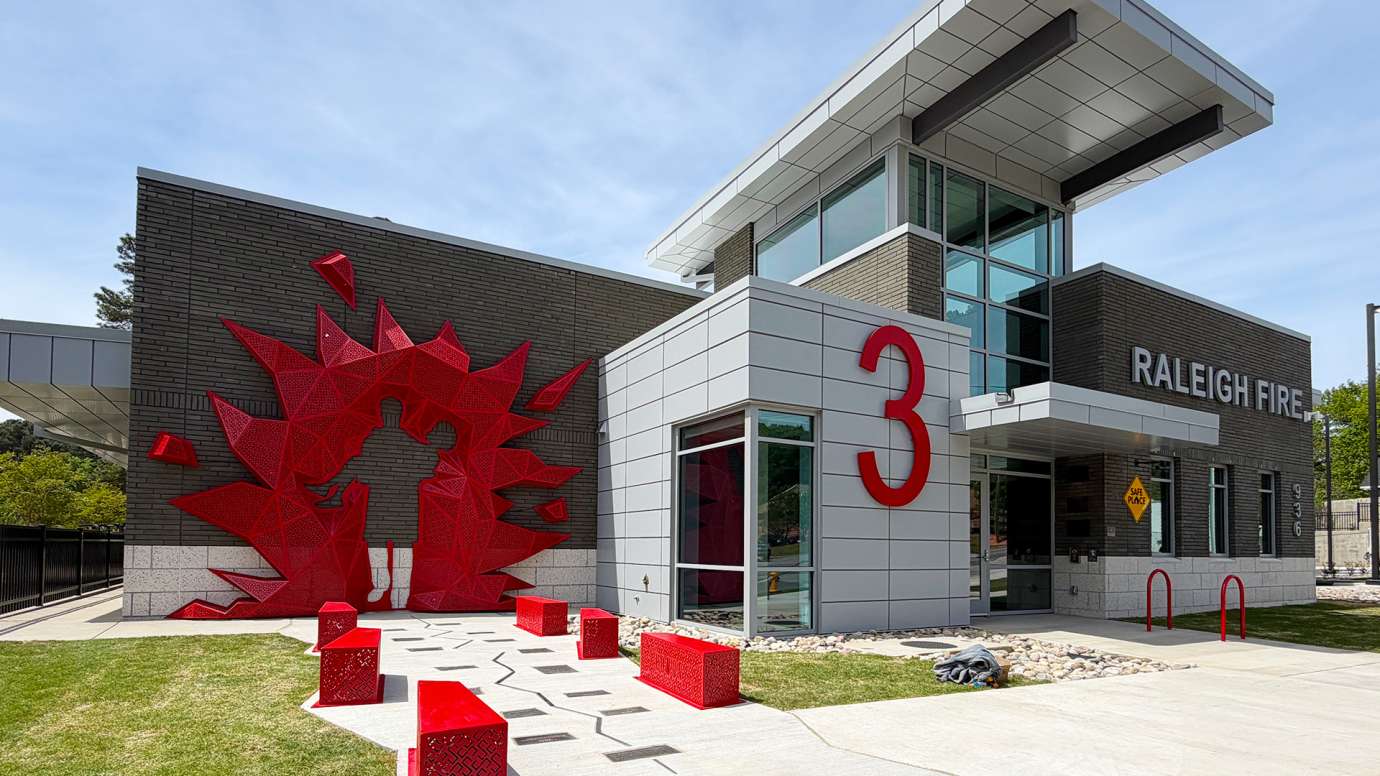 A red metal sculpture with the outline of a firefighter rescuing a woman is on the wall of a fire station building.