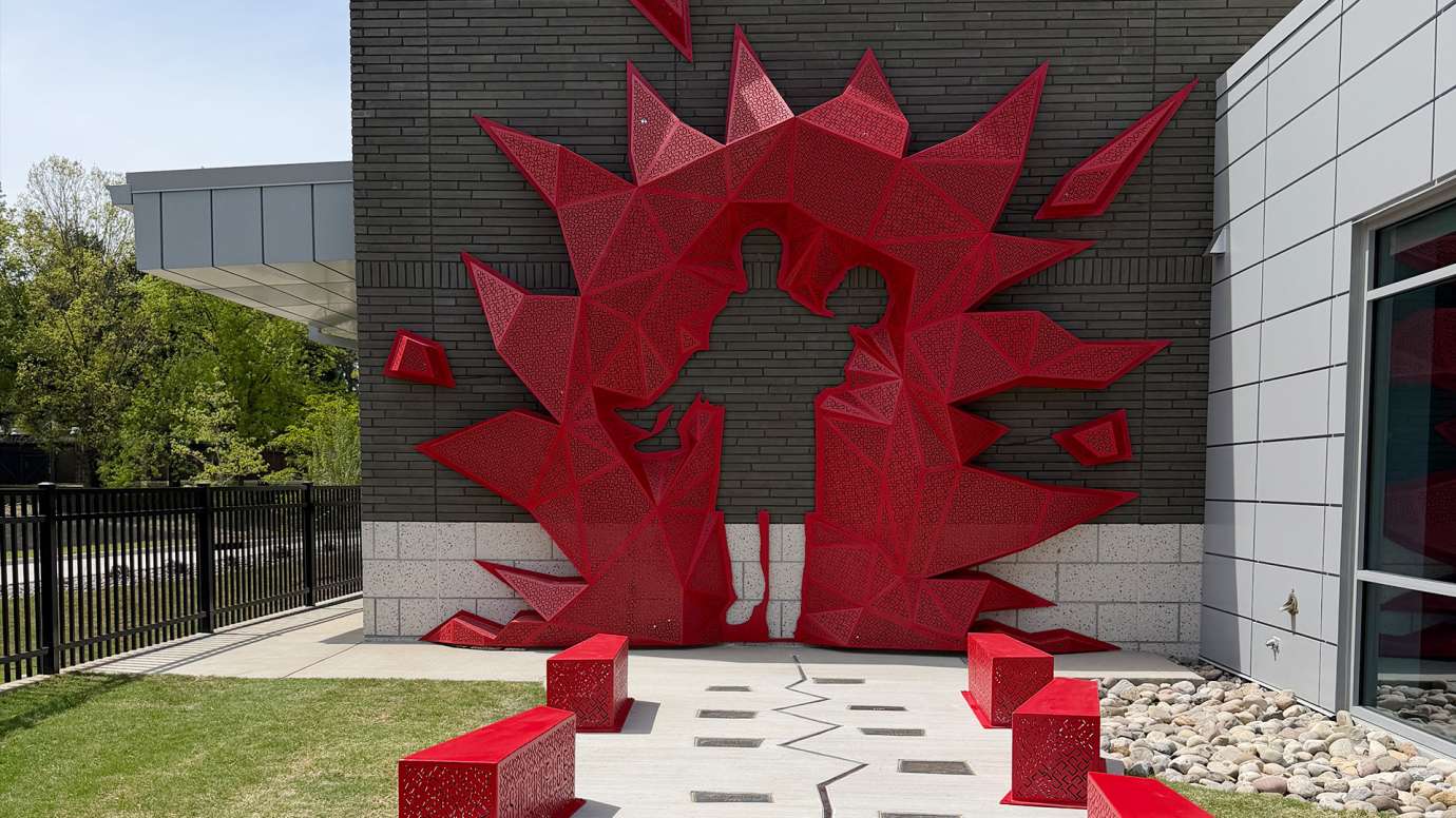 An image of a red metal sculpture with the outline of a firefighter carrying a person is attached to a brick wall. There are red benches and plaques in front of it.