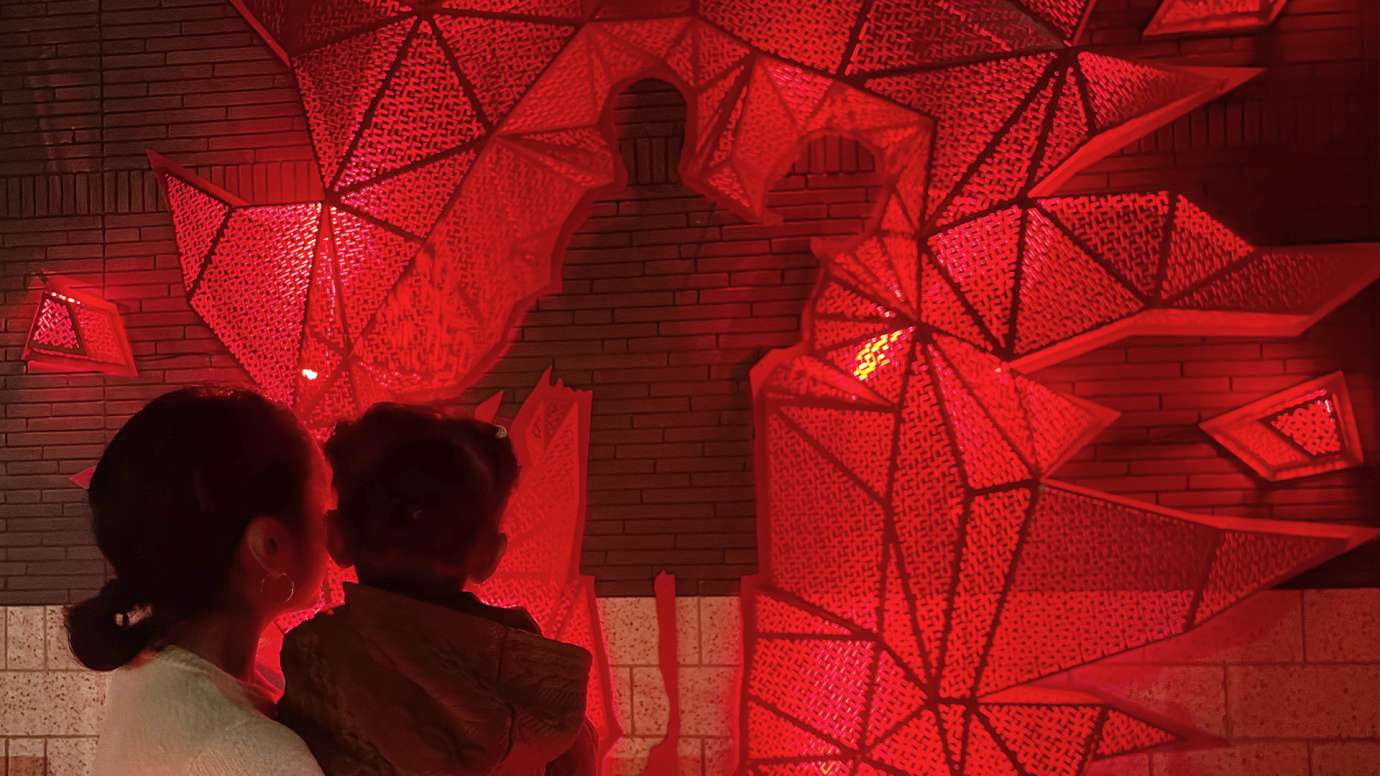 A woman and child look closely at the artwork which is glowing red.