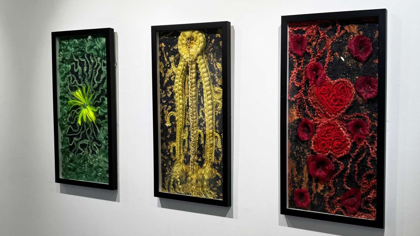 Three mixed media works in black frames by Chris-Annia Dobson, the far left work in shades of green, the middle piece in shades of yellow, and the work on the right in shades of red
