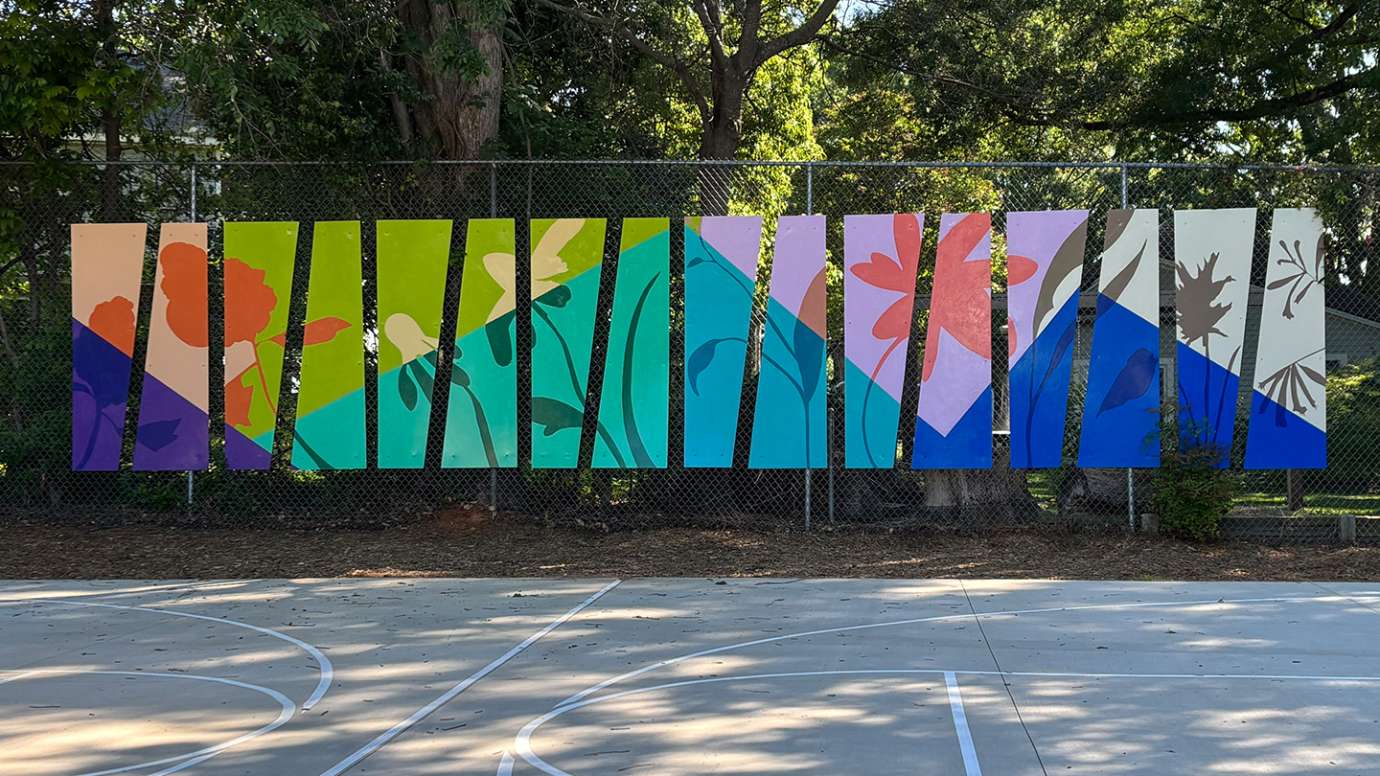 Sixteen brightly colored metal panels are hanging on a chain link fence. They are painted with silhouettes of native flowers.