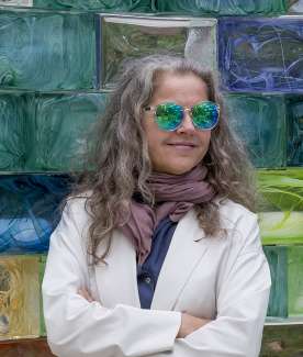 A woman leans against a wall of colorful blocks wearing sunglasses.