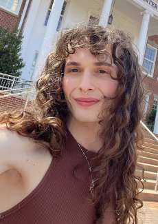 The artist smiles at the camera in a brown shirt with long curly hair.