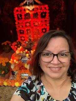 Woman in glasses smiling in front of a bright red ofrenda display. 