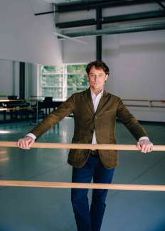Man standing at a ballet barre with a green textured blazer and jeans. 