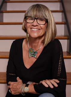 Headshot of Deb Karash - a woman smiling at the camera with black framed glasses and a bob, wearing a large metal necklace with turquoise colored flowers and a black v-neck shirt