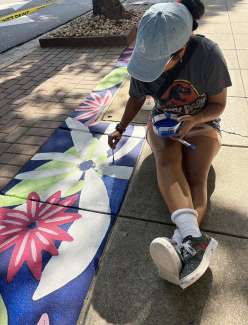 A woman in a baseball hat is sitting on a sidewalk painting a floral design.
